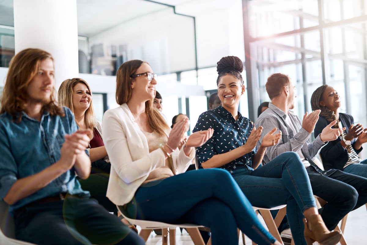 A group of people sitting in chairs, attentively watching and clapping in a bright, modern interior as they eagerly await the announcement of choosing a keynote speaker.