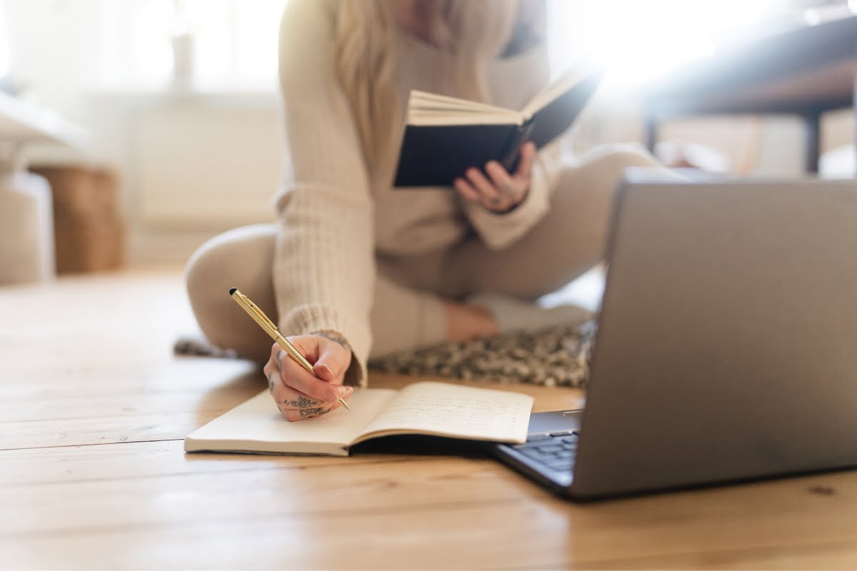 A person sits on the floor with legs crossed, writing in a notebook while holding a book, with an open laptop in front of them, seemingly engrossed in the task of choosing a keynote speaker.