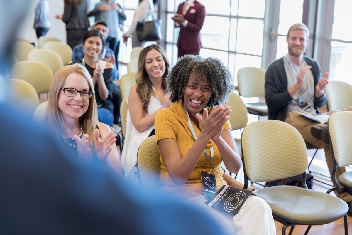 Audience members seated in rows clap and smile, showing positive reactions during the presentation on choosing a keynote speaker in a brightly lit room.