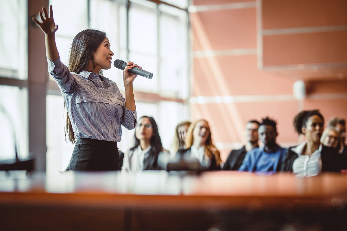 A woman speaks into a microphone while gesturing with her hand, embodying the essence of choosing a keynote speaker, in front of an audience seated in blurred background.