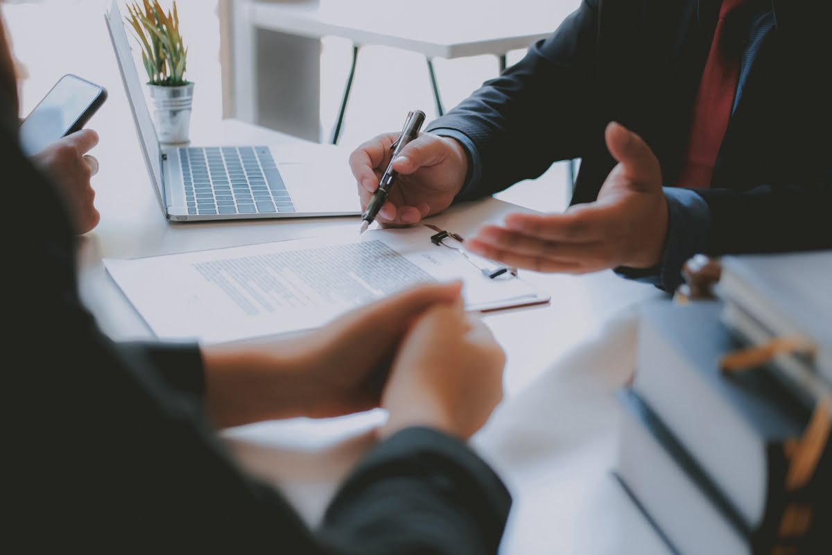 Two people are sitting at a desk discussing a document. One person is holding a pen and pointing at the document, possibly choosing a keynote speaker, while the other listens with hands clasped. A laptop and a stack of books are on the desk.