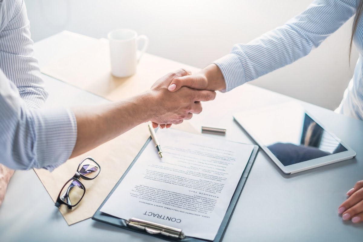 Two individuals shake hands over a signed contract on a clipboard, finalizing their choice for a keynote speaker. Nearby are eyeglasses, a tablet, a pen, and a coffee mug on the desk.