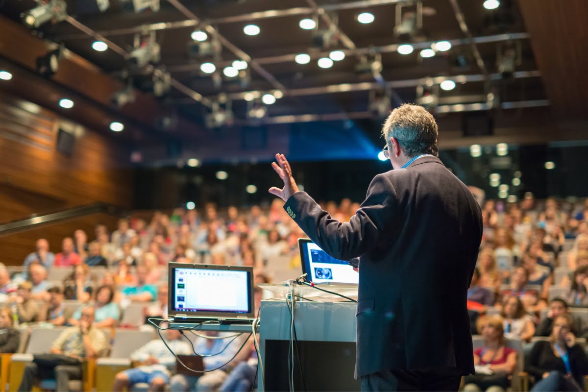 A speaker, chosen as the keynote speaker, addresses a large audience in an auditorium, with a laptop and screen in front of him displaying a presentation.