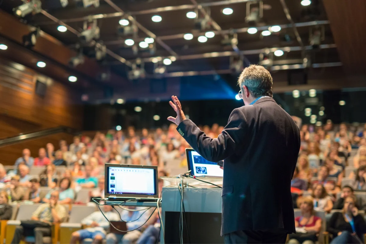 A speaker, chosen as the keynote speaker, addresses a large audience in an auditorium, with a laptop and screen in front of him displaying a presentation.