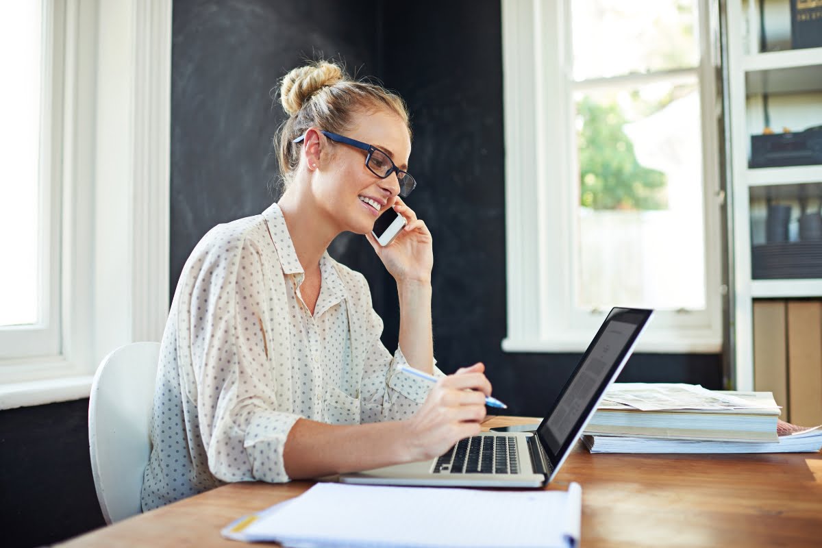 A woman wearing glasses and a polka dot shirt is talking on the phone and working on a laptop at a desk in a sunlit room, diligently choosing a keynote speaker for an upcoming event.