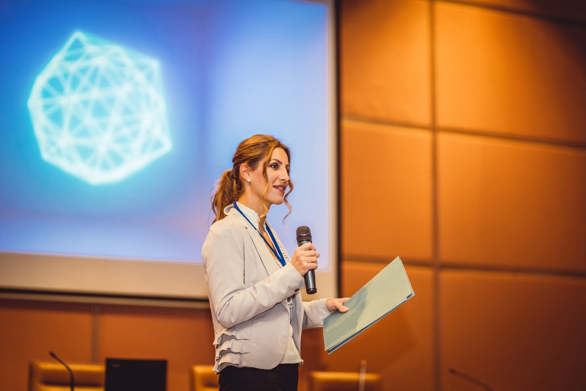 A woman in a light blue blouse, chosen as the keynote speaker, holds a microphone and a folder, speaking at a podium with a geometric design projected on the screen behind her.