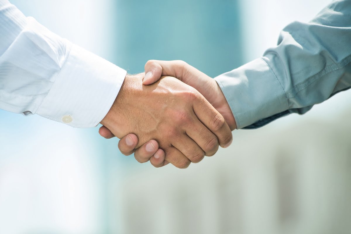 Two people wearing dress shirts shake hands against a blurred outdoor background, exemplifying effective communication strategies.