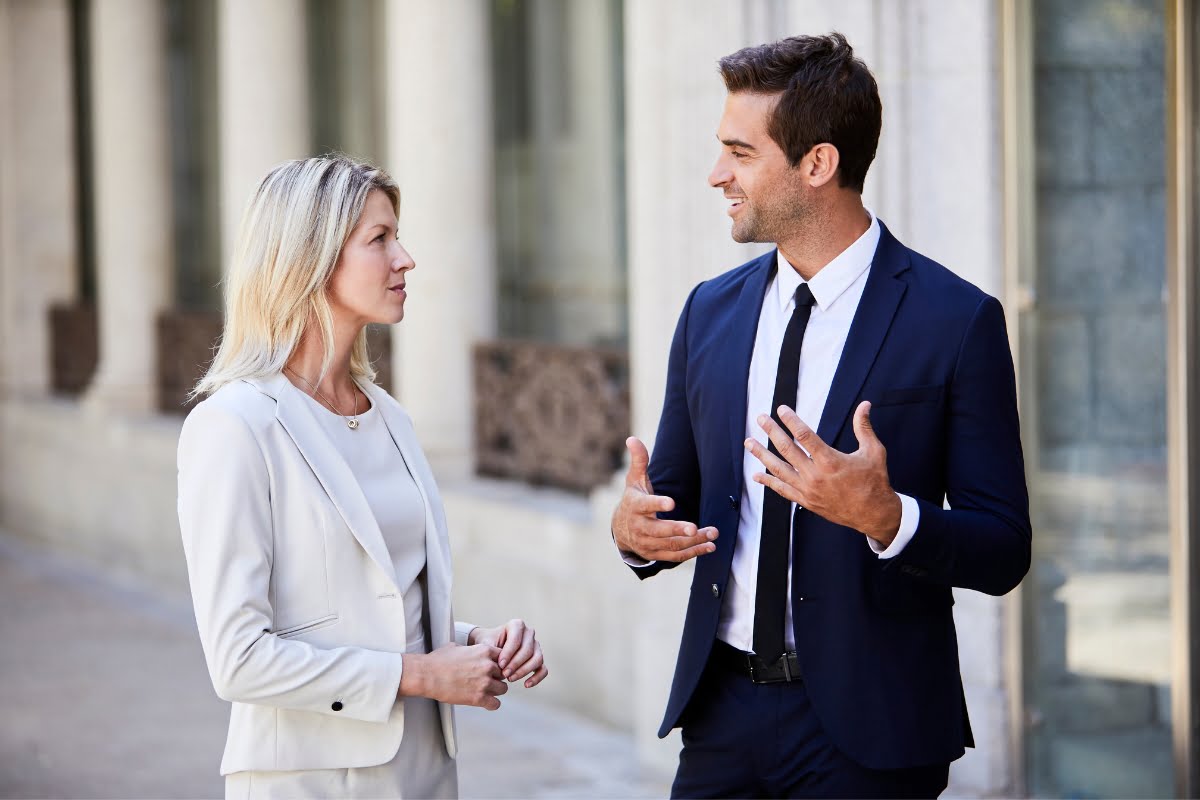 A man and a woman in business attire discuss effective communication strategies outside near a building.