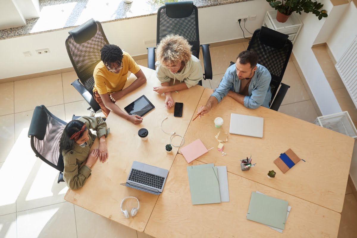 Four people sit around a table with laptops, tablets, and notebooks, engaged in a discussion about effective communication strategies in a bright office setting.