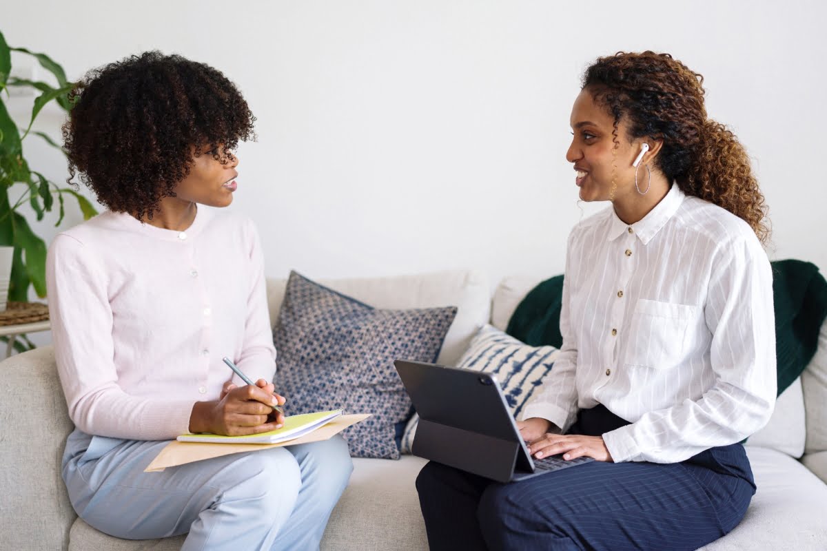 Two women sitting on a sofa, one taking notes on a clipboard while the other uses a tablet, appear to be discussing effective communication strategies.