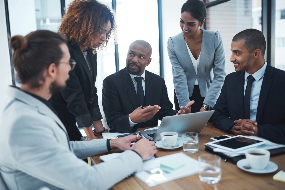 A group of five business professionals having a discussion around a conference table with laptops, tablets, and coffee cups, focusing on effective communication strategies.