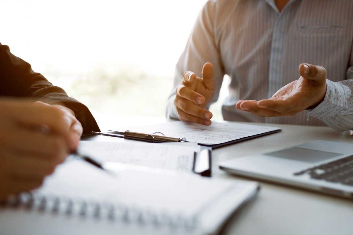 Two people are having a discussion at a desk with documents, a laptop, and a notepad; one person's hands are gesturing while the other is holding a pen, showcasing effective communication strategies.