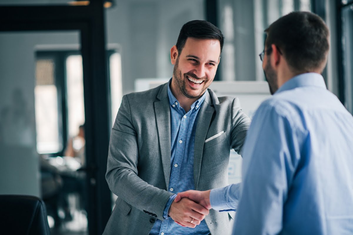 Two men are shaking hands and smiling in a professional setting, with an office environment in the background. One is wearing a gray blazer, and the other a light blue shirt. Their interaction exemplifies effective communication strategies, fostering mutual respect and understanding.