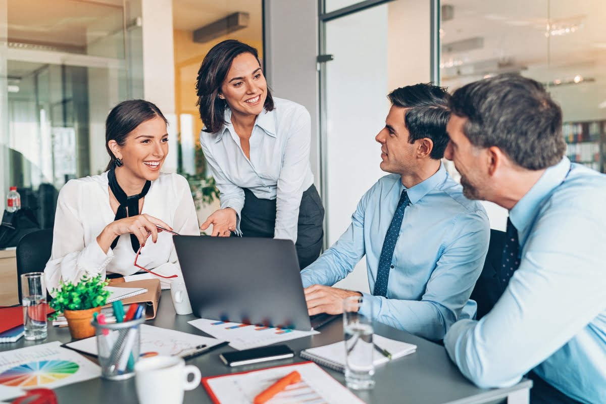 Four colleagues in business attire have a discussion around a laptop in a modern office setting, focusing on effective communication strategies. Papers, charts, and office supplies are on the table. The mood appears collaborative and positive.