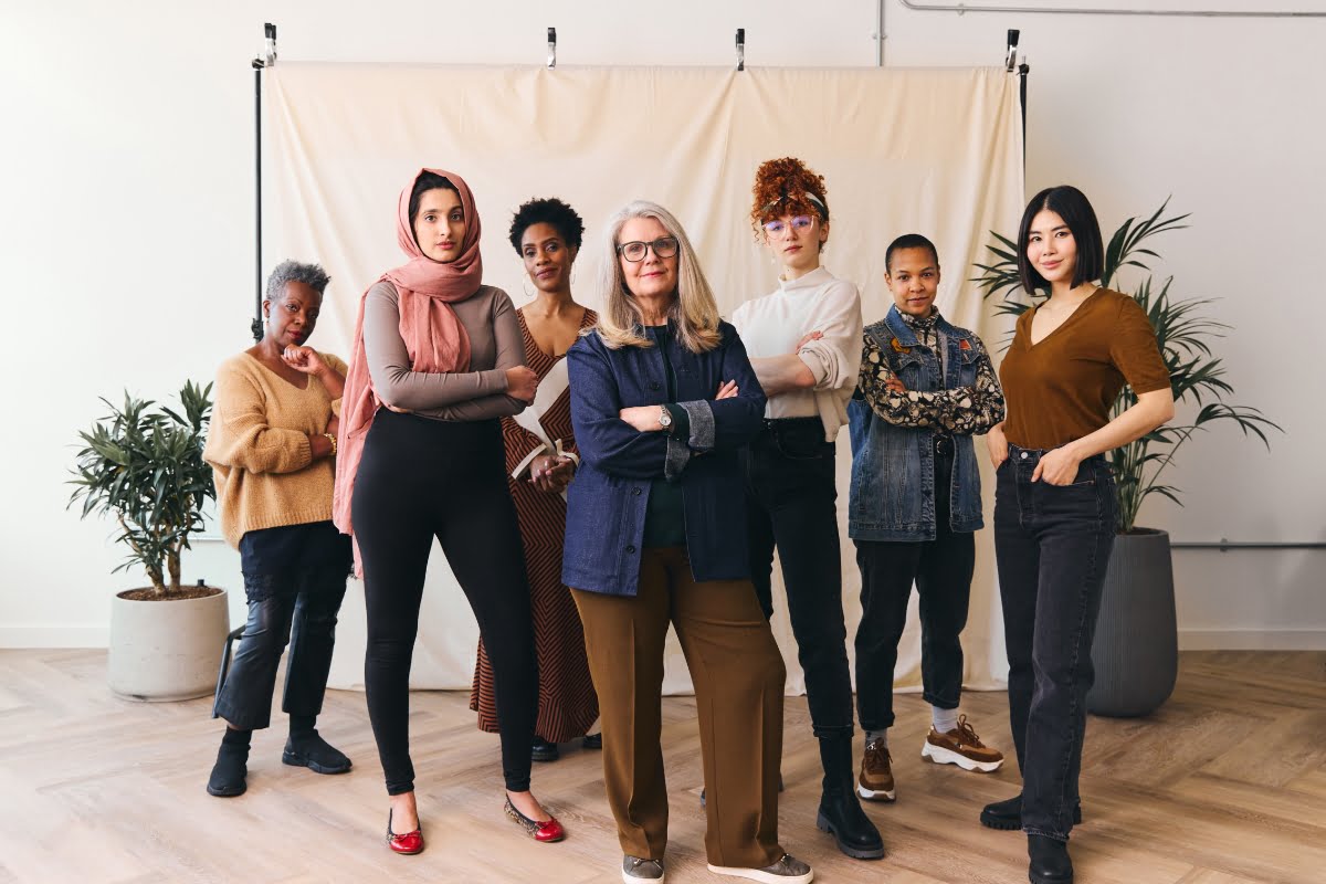 A diverse group of seven women, each standing confidently with their arms crossed, in front of a beige backdrop. They symbolize strength and unity in addressing menopause in the workplace. Lush plants are placed on both sides of the group, adding a touch of nature to the scene.