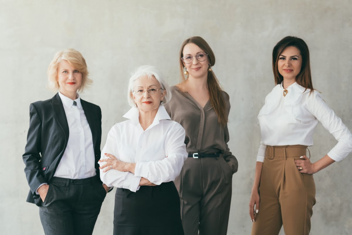 A group of four women standing together in front of a plain background, dressed in business casual attire, showcasing solidarity and expertise in addressing menopause in the workplace.