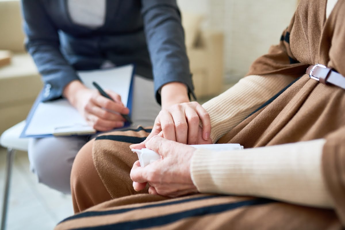 A person in a gray blazer takes notes while another individual in a brown robe, with visible hands, holds a tissue and receives a comforting hand gesture, highlighting the importance of addressing menopause in the workplace.