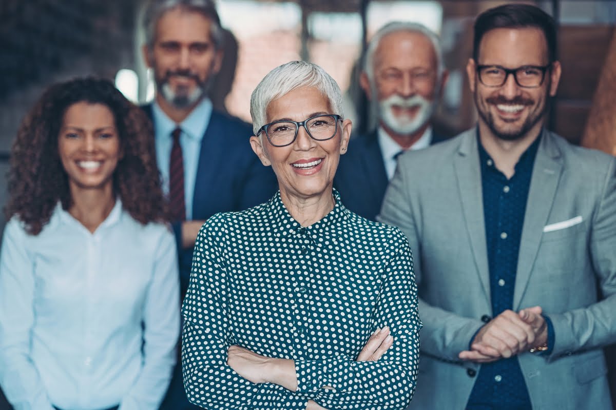 A group of five professionals, including two women and three men of varying ages, stand together smiling. The woman in front has short white hair and glasses, possibly symbolizing her experience and resilience during menopause in the workplace.