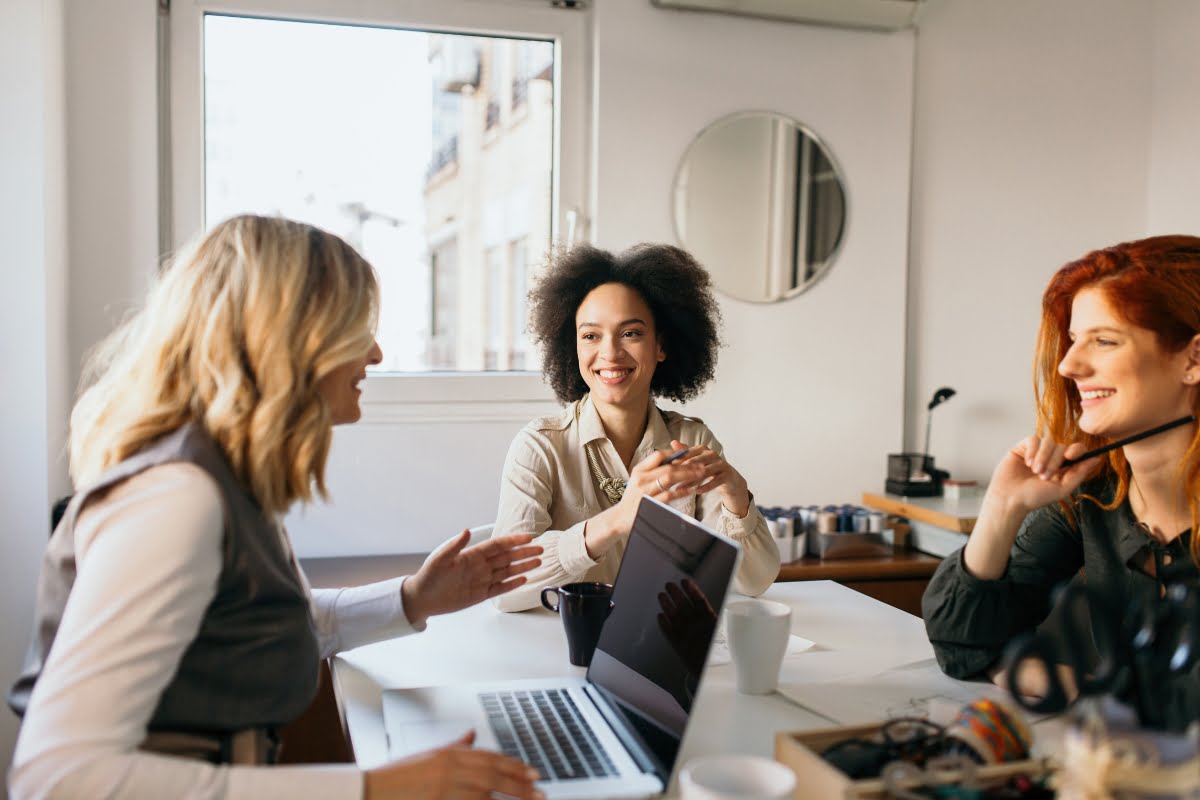 Three women are sitting at a table in an office. One is working on a laptop, while the other two are engaged in conversation, smiling and discussing menopause in the workplace.