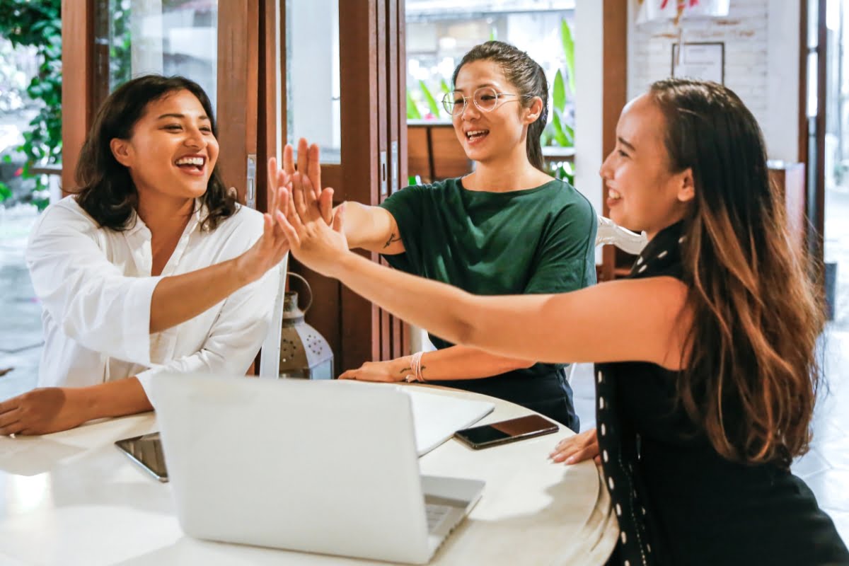 Three women sitting at a table high-five each other near an open laptop, celebrating a positive and animated conversation about menopause in the workplace in a casual indoor setting.