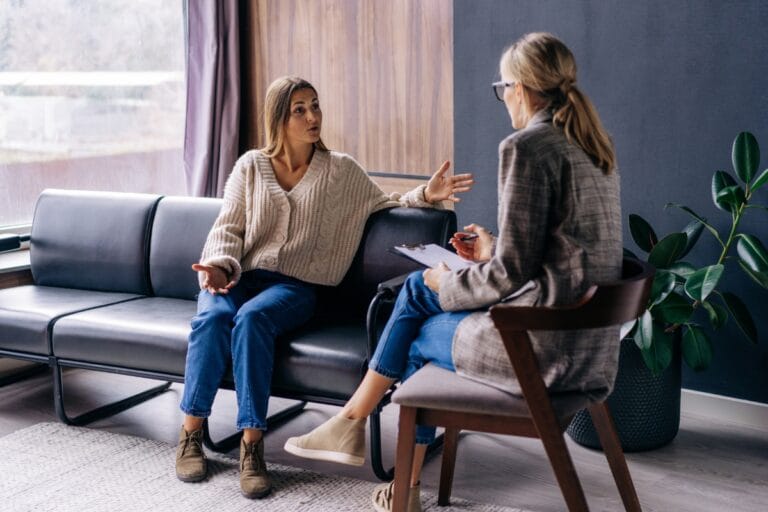 Two women sit across from each other in a serene living room, engaging in mental health counseling. One gestures expressively, while the other takes notes on a clipboard. A potted plant and large window provide a calming backdrop to their earnest exchange.