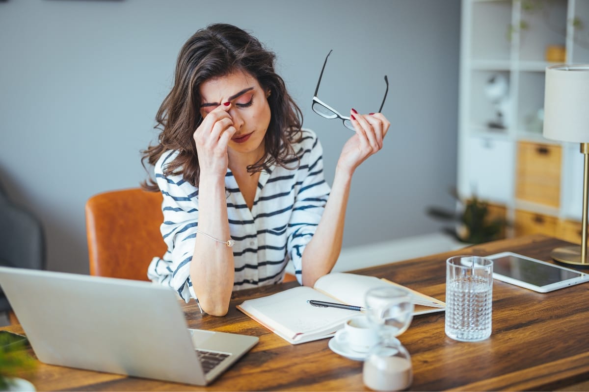 A woman in a striped shirt sits at a desk, holding her glasses and rubbing her eyes, reflecting on her mental health counseling session. In front of her are a laptop, notebook, coffee cup, and a glass of water.