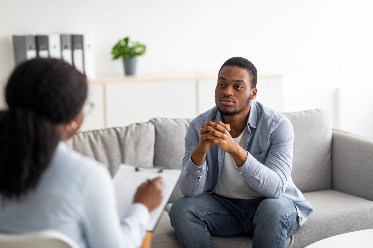 A man sits on a gray sofa, hands clasped, attentively listening in a mental health counseling session as the woman with a clipboard guides him through their office discussion.