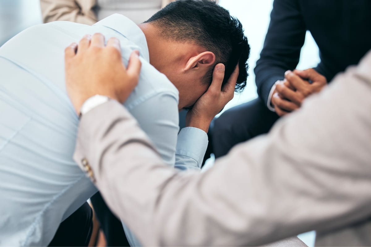 A person leans forward, resting their head in their hand, while two others offer support by placing hands on their back and shoulder, symbolizing the comforting embrace of mental health counseling.