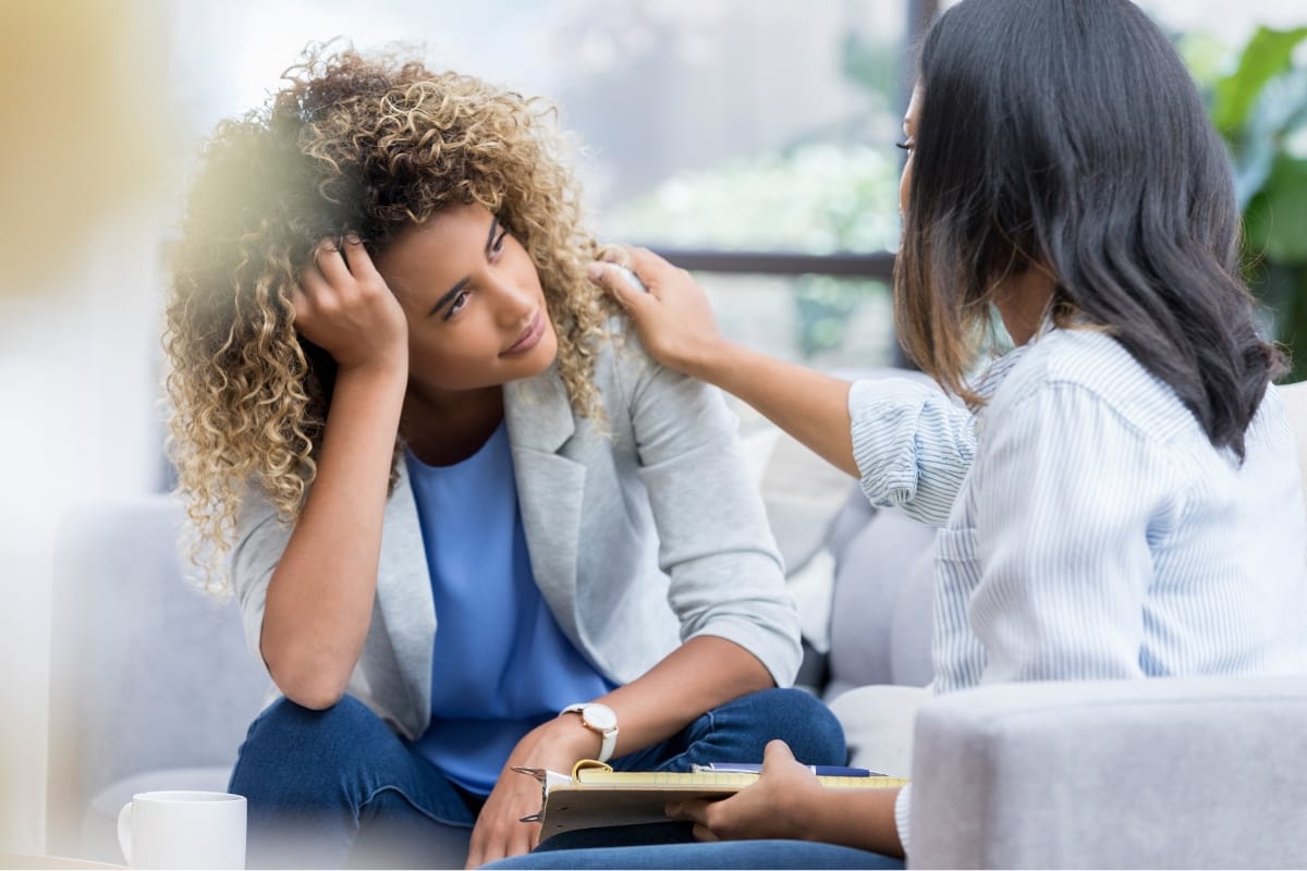 Two women are sitting on a couch; one appears to be offering comfort through what seems like an informal session of mental health counseling, as the other looks visibly distressed.