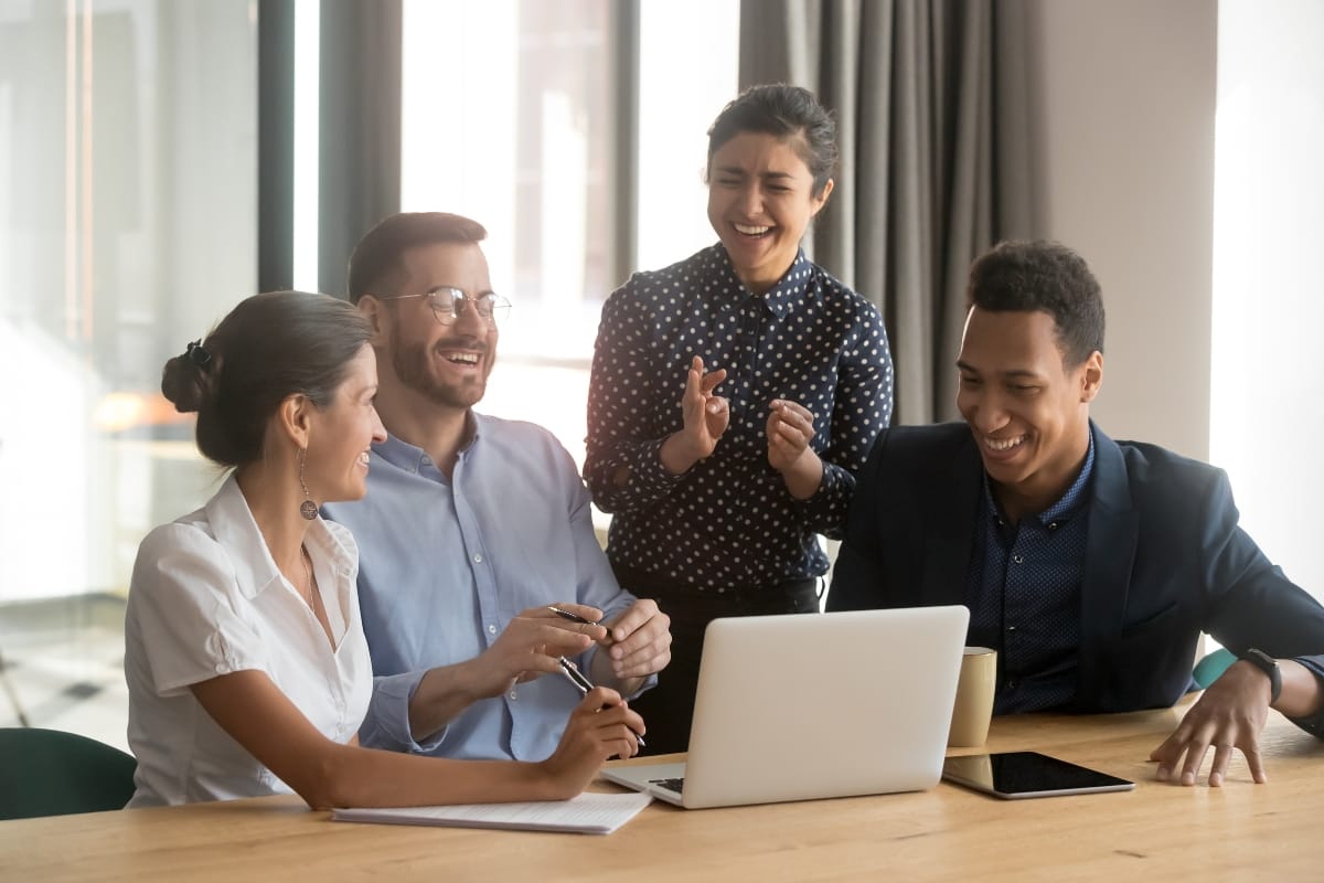 Four people are gathered around a laptop, smiling and talking in a well-lit room. Their engaging discussion centers on mental health counseling, fostering an atmosphere of understanding and support.