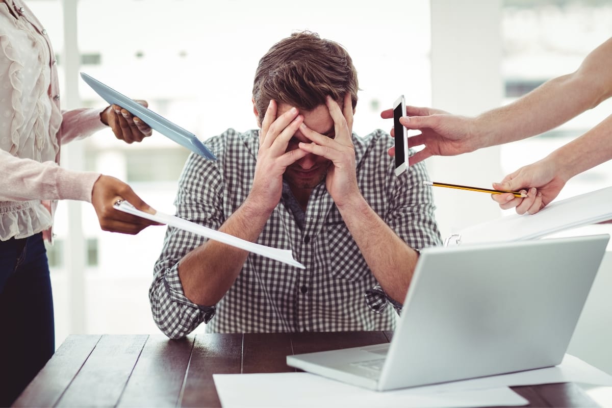 Person sitting at a desk, holding their head in frustration, surrounded by multiple hands with a phone, documents, and a pen—a chaotic scene that underscores the need for mental health counseling in such a stressful work environment.