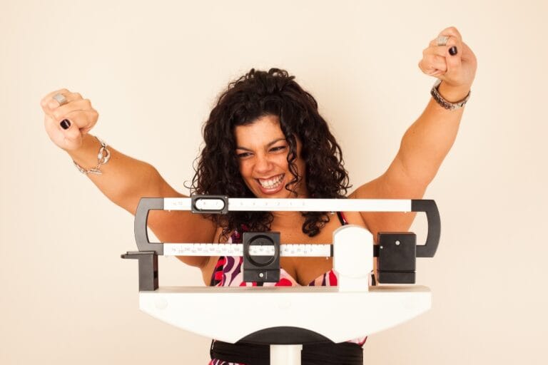 Woman smiling with raised fists while standing on a scale, embracing her weight loss solutions.