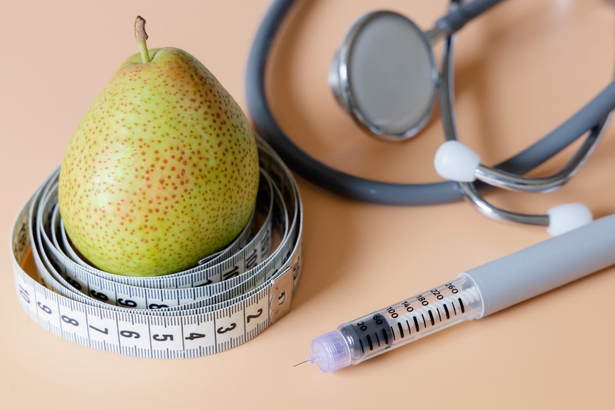 A pear wrapped in a measuring tape next to a syringe and a stethoscope on a peach-colored surface illustrates innovative weight loss solutions.