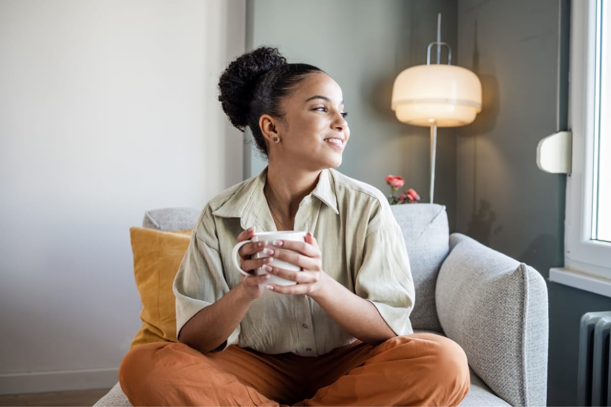 A person sits cross-legged on a couch, holding a mug and softly smiling at the window, seemingly contemplating their weight loss solutions. A lamp and a cushion rest in the background, adding warmth to the serene scene.