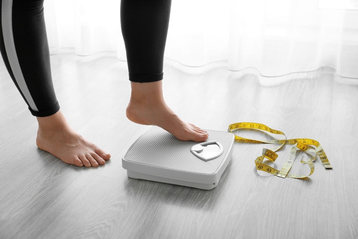 A person steps onto a digital scale near a yellow measuring tape on a wooden floor, exploring effective weight loss solutions.