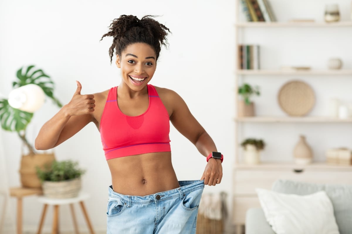 A woman in a red sports bra holds out the waistband of oversized jeans, smiling and giving a thumbs up in a bright living room, celebrating her incredible weight loss solution.