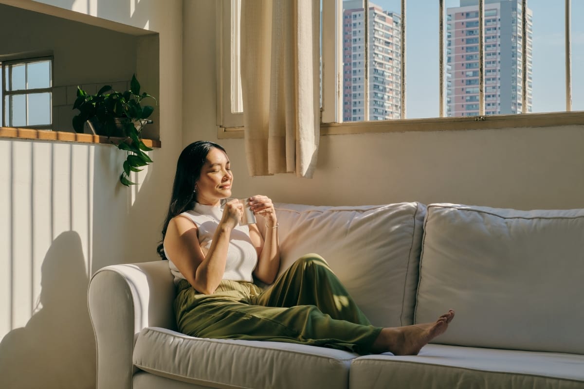 A woman sits on a white couch by the window, basking in sunlight. Dressed in a white top and green pants, she contemplates her weight loss solutions while two tall buildings stand majestically outside.