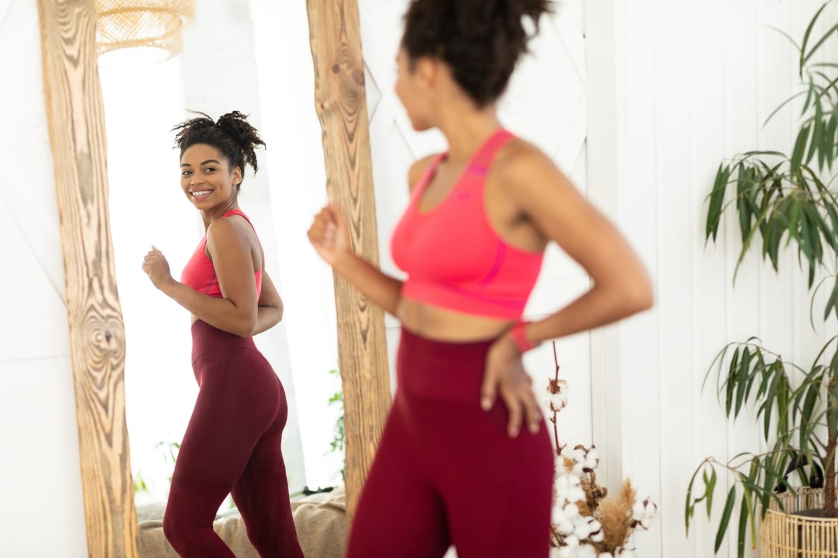 A woman in a pink sports bra and maroon leggings smiles at her reflection in the mirror, proud of her progress and thanking her commitment to effective weight loss solutions.