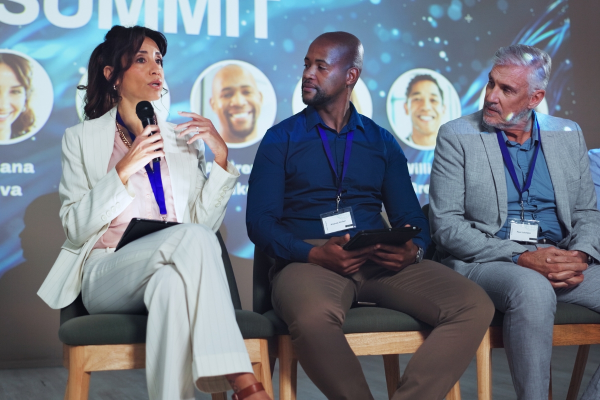 Three keynote speakers for business events are seated on stage, engaging in a panel discussion. A woman on the left speaks into a microphone while the two men listen attentively. Behind them, a digital screen displays relevant images and text.