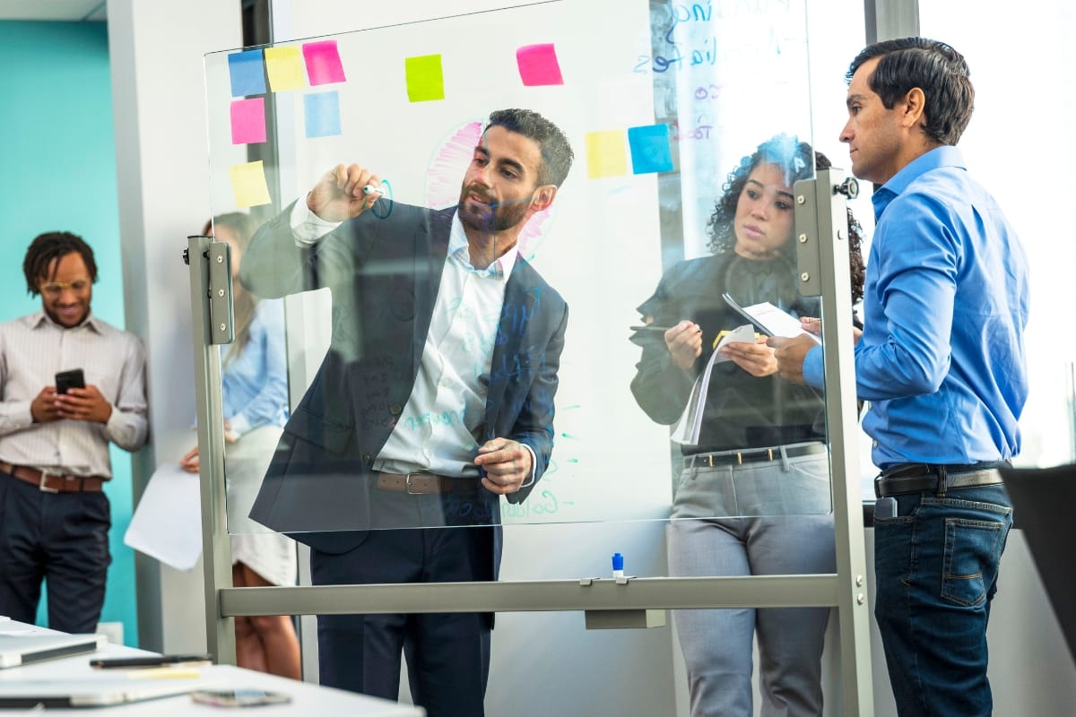 A man writes on a transparent board with markers, reminiscent of keynote speakers for business events, while three colleagues look on—two holding notepads and one using a smartphone. Bright sticky notes dot the board.