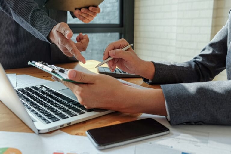 Two professionals engage in a problem-solving discussion at a desk, surrounded by documents, a laptop, and a smartphone. One points at a clipboard while the other holds a pen, illustrating strategic problem solving for leaders amid scattered papers and a calculator.