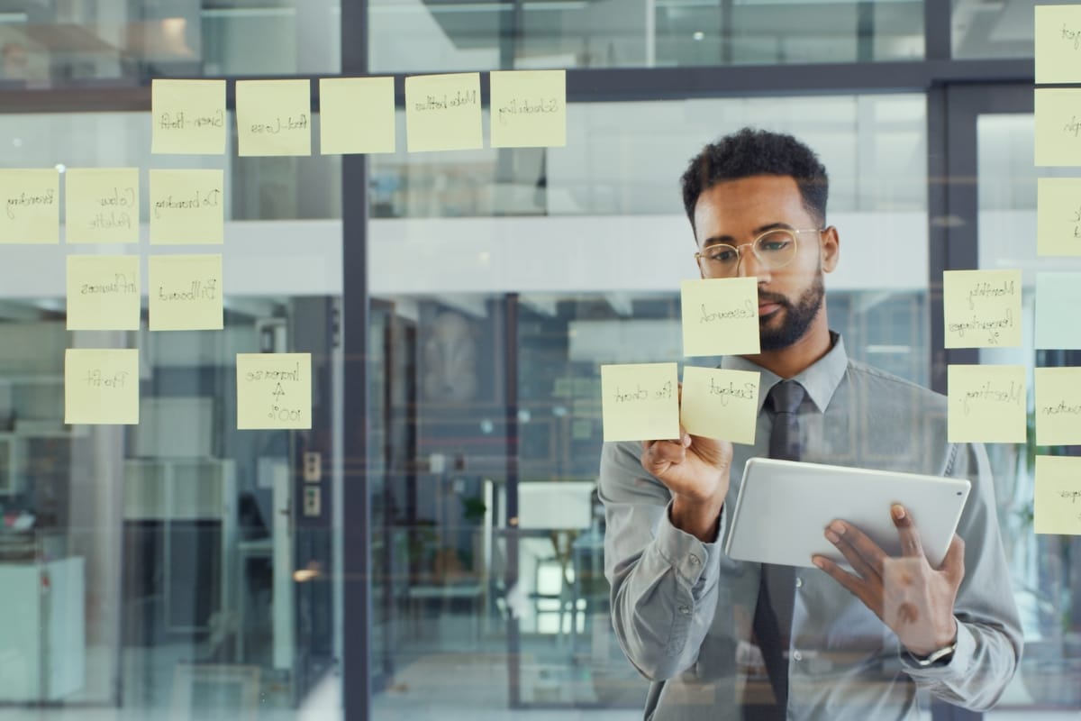 A person holding a tablet stands in front of a glass wall covered with sticky notes, embodying the spirit of problem solving for leaders in an office setting.