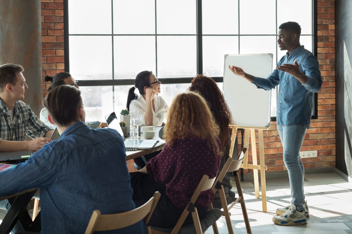 A person stands near a whiteboard, giving a presentation on problem solving for leaders to a group seated around a table in a well-lit room with large windows.