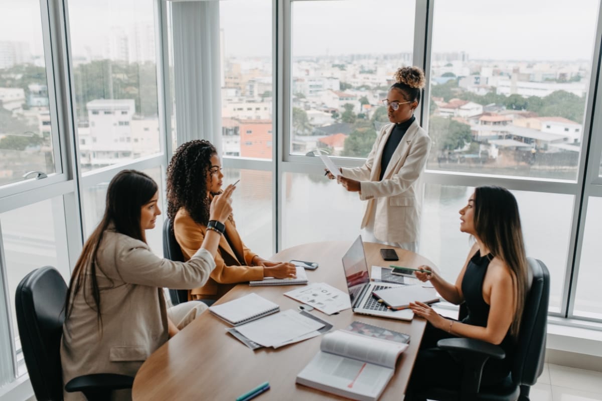 Four leaders in a meeting room with large windows are engaged in dynamic problem solving, as they sit and stand around a table laden with laptops, papers, and notebooks. One person is speaking passionately while holding a document, guiding the discussion forward.