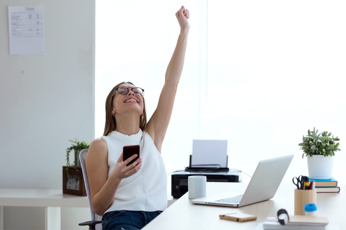 A woman sits at a desk, smartphone in hand and other arm raised in celebration, embodying problem-solving for leaders. Nearby, a laptop and coffee mug hint at her successful day.