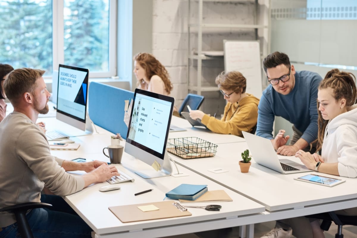 People working collaboratively in a modern office, using computers and tablets, focus on problem solving for leaders. A whiteboard and large windows are visible in the background.