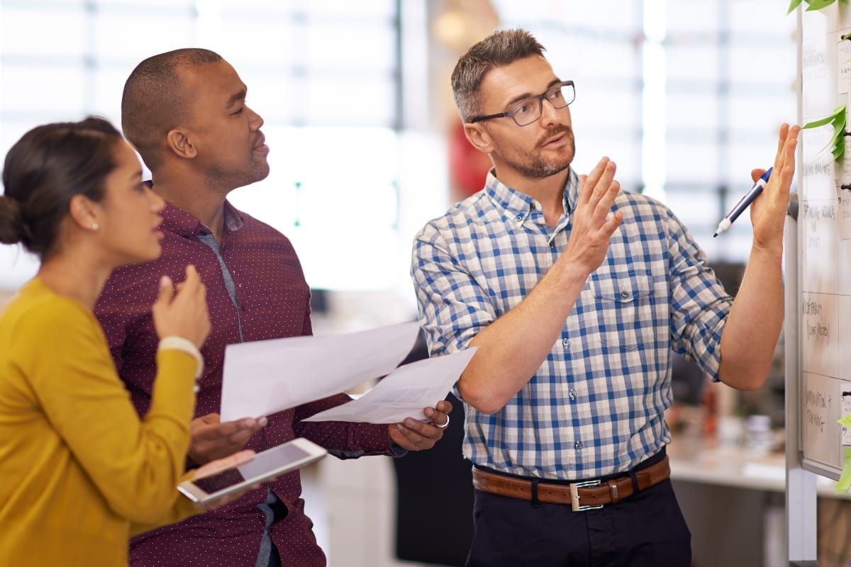 Three people engage in a dynamic session at the whiteboard; one gestures with a pen, emphasizing key points of problem-solving for leaders, while the others hold papers and listen attentively.