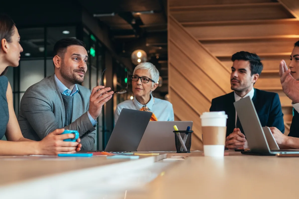 A mastermind group of five people sits in a meeting room, engaged in lively discussion around a table filled with laptops and drinks.