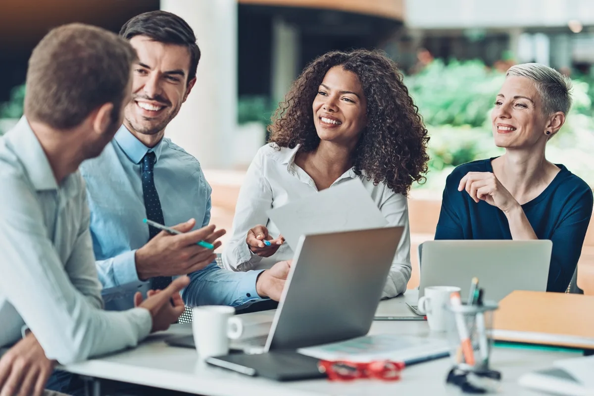 Four people sitting at a table with laptops, papers, and cups, are engaged in a lively mastermind group discussion, sharing insights and smiling.