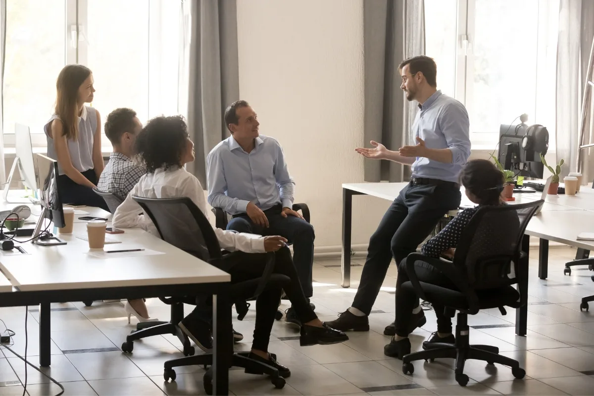 A group of six people in a brightly lit office room are seated and standing near computers, engaged in a discussion on employee productivity, blending mindful leadership with traditional styles for optimal outcomes.
